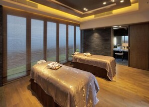 A modern spa room with two massage tables, a large window with blinds, a dark textured wall, and a small bathroom with a sink and mirror. - The Royal Garden Kowloon East (Sai Kung)