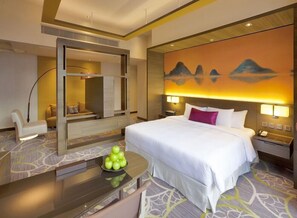 Une chambre d’hôtel avec un lit, un bureau sur lequel se trouve un bol de pommes, une chaise, un grand miroir et un mur orné d’un tableau représentant une montagne.