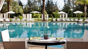 Mesa à beira da piscina com copos e cardápio, rodeada por espreguiçadeiras e guarda-sóis, com piscina e vegetação ao fundo.
