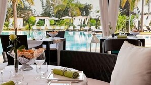 Mesa de jantar com vista para uma piscina, área de estar externa e paisagem tropical.