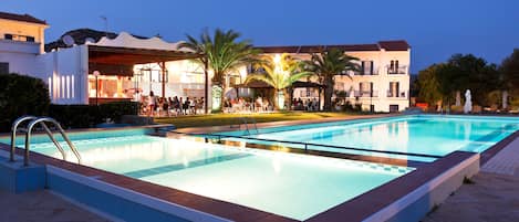 A swimming pool with a tiled deck, surrounded by palm trees and a building with multiple balconies.