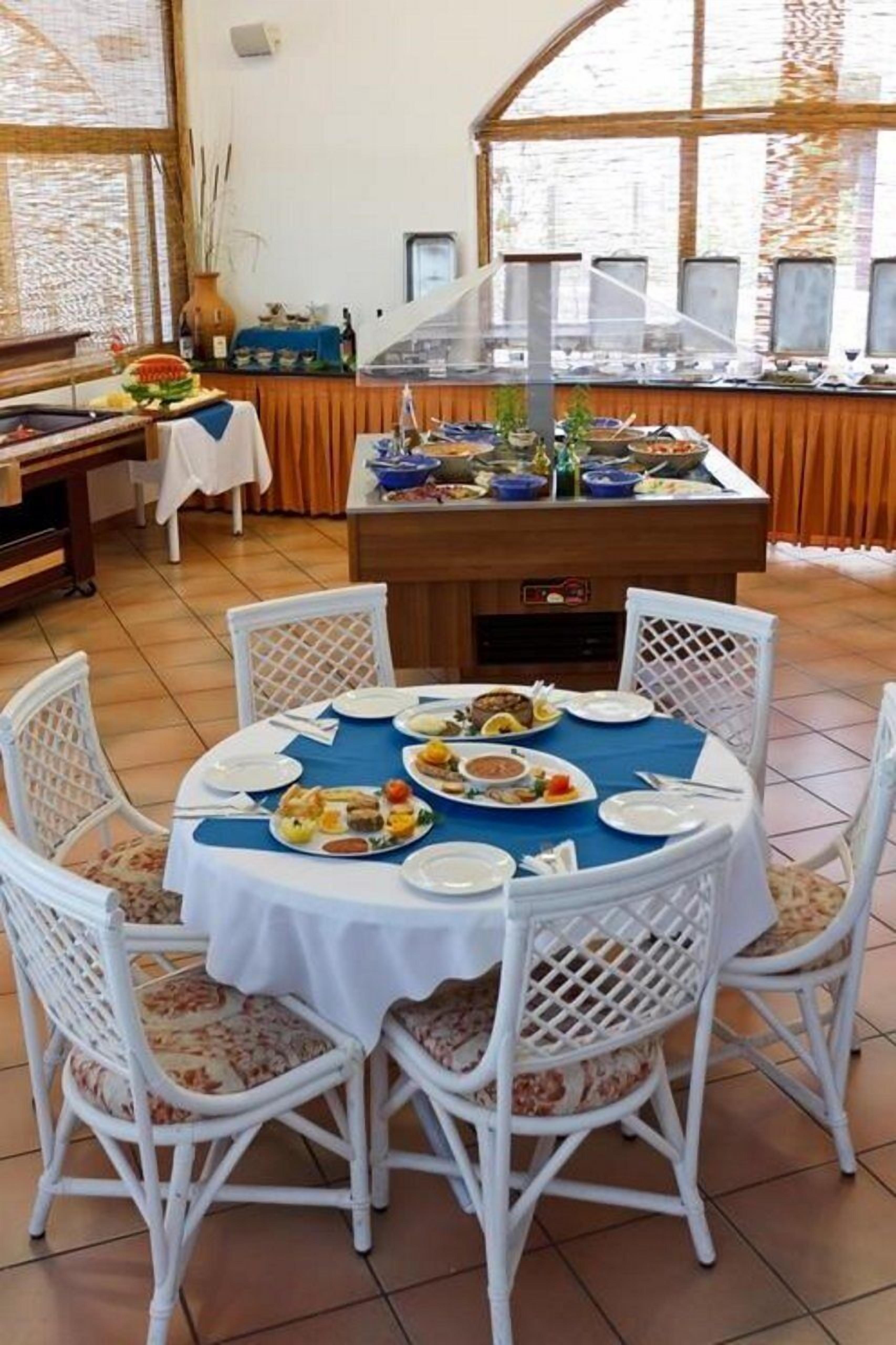 A dining area with a round table set for four, surrounded by white wicker chairs. A buffet counter with various dishes is visible in the background.