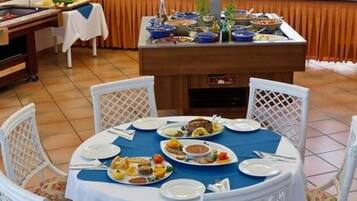 A dining area with a round table set for four, surrounded by white wicker chairs. A buffet counter with various dishes is visible in the background.