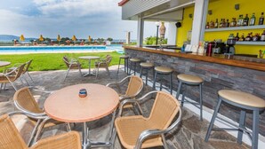 Un espace bar extérieur avec des fauteuils en osier et une table ronde, offrant une vue sur une piscine et une plage.