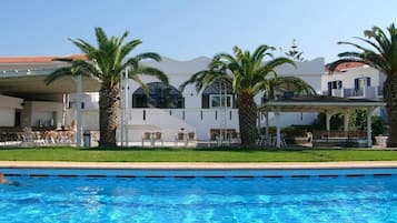 Uitzicht op een zwembad van een resort, met palmbomen, een overdekte zithoek en een gebouw met een witte gevel en een dak van rode tegels.