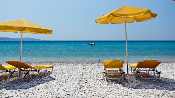 Une plage avec des parasols jaunes et des chaises longues, une plage de galets et un bateau sur l’eau.