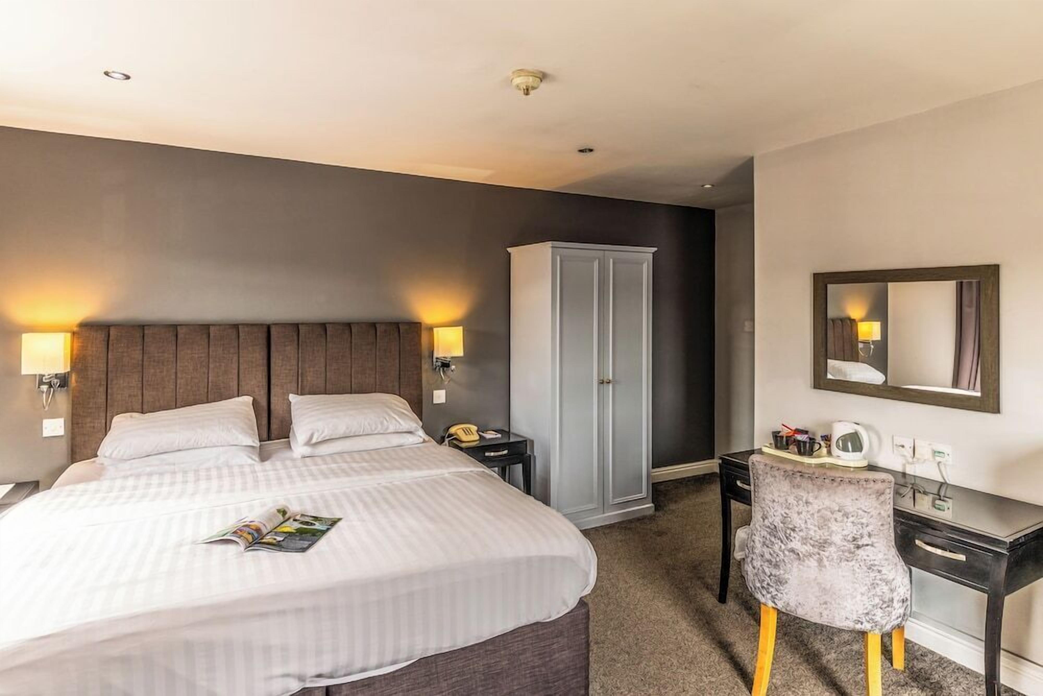 A hotel room with a large bed, bedside lamps, a wardrobe, a desk with a chair, and a mirror on the wall.