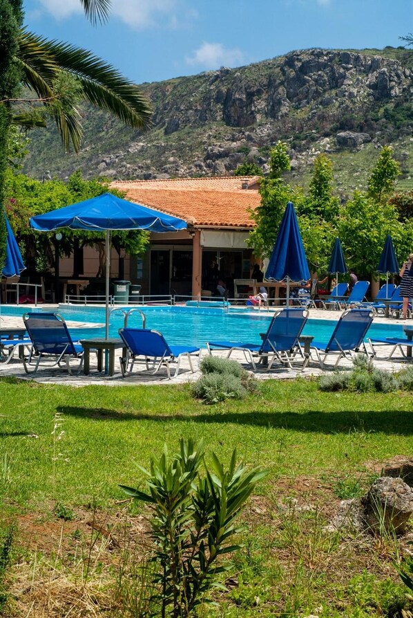 Zona de piscina con sillones reclinables azules y sombrillas, rodeada de vegetación y con una montaña al fondo.