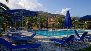 A poolside area with blue lounge chairs and umbrellas, a clear sky, and a mountain in the background. - Efrosini Village (Kefalonia)
