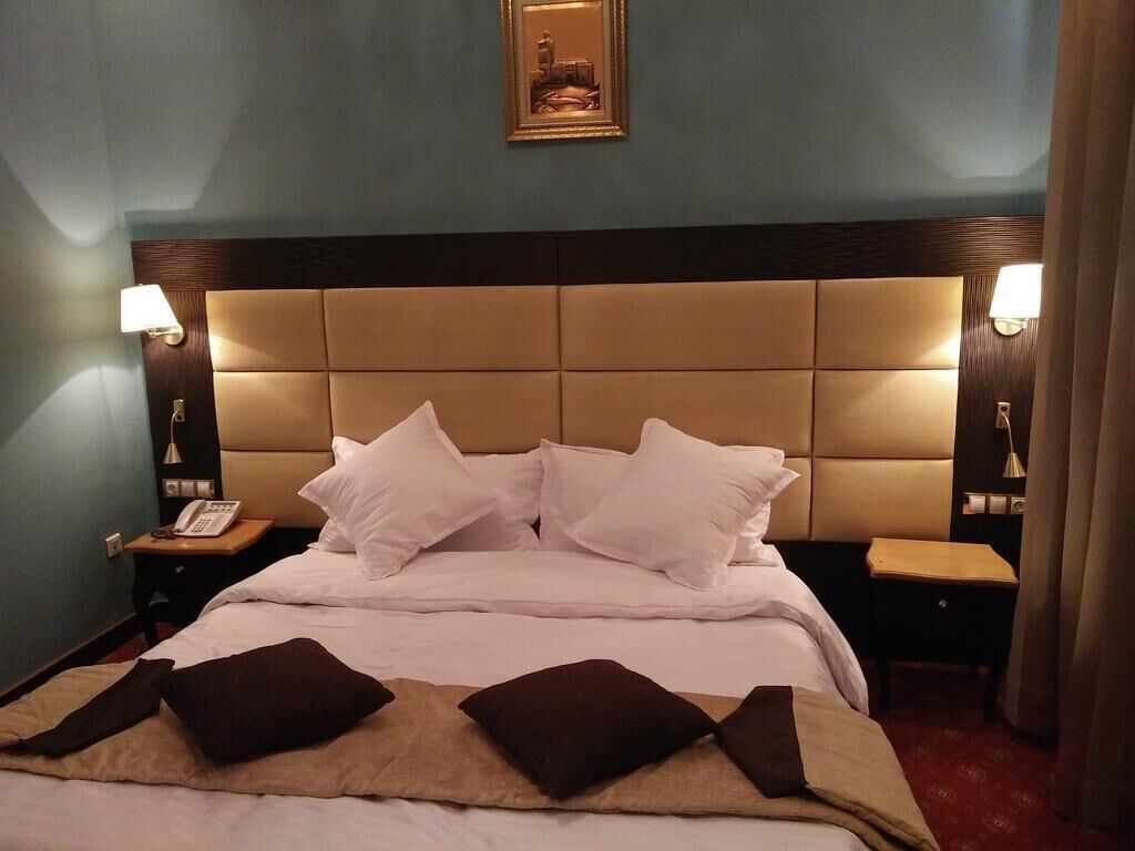 Une chambre d’hôtel avec un grand lit, deux tables de chevet, une petite table, un téléphone fixé au mur et un tableau encadré au-dessus du lit.