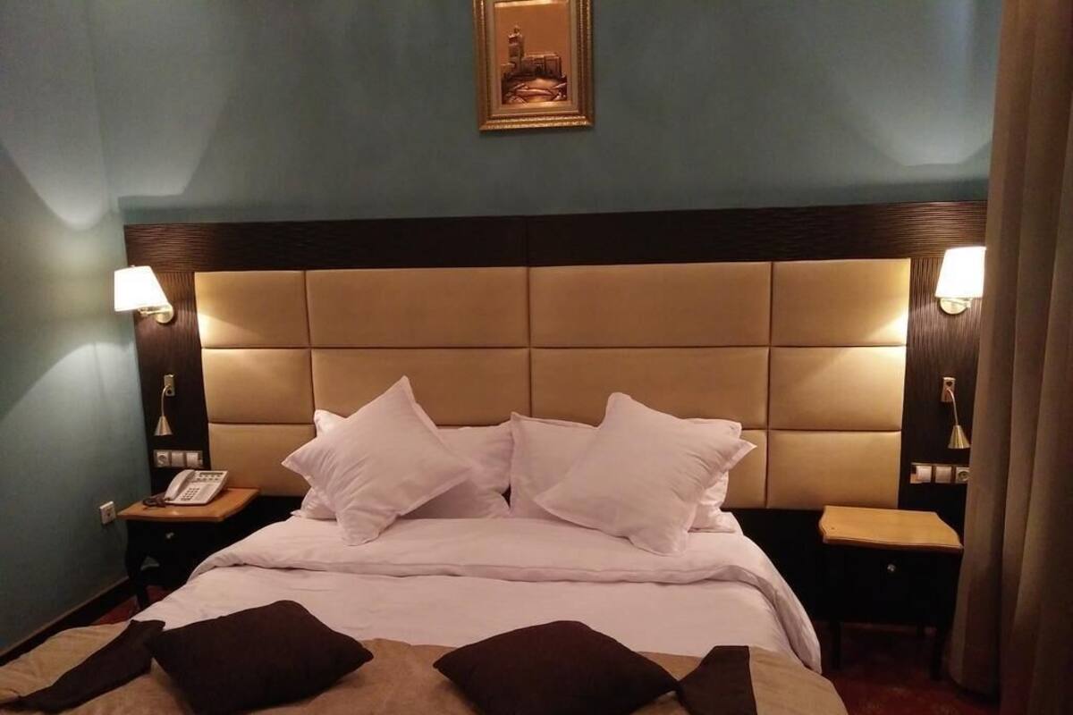 Une chambre d’hôtel avec un grand lit, deux tables de chevet, une petite table, un téléphone fixé au mur et un tableau encadré au-dessus du lit.