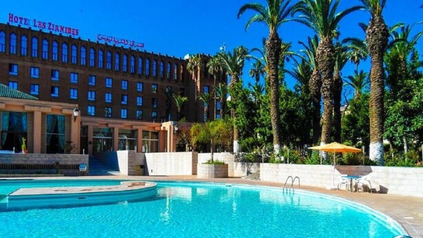 A hotel with a swimming pool, palm trees, and a clear blue sky. - Hotel Les Zianides (Tlemcen)