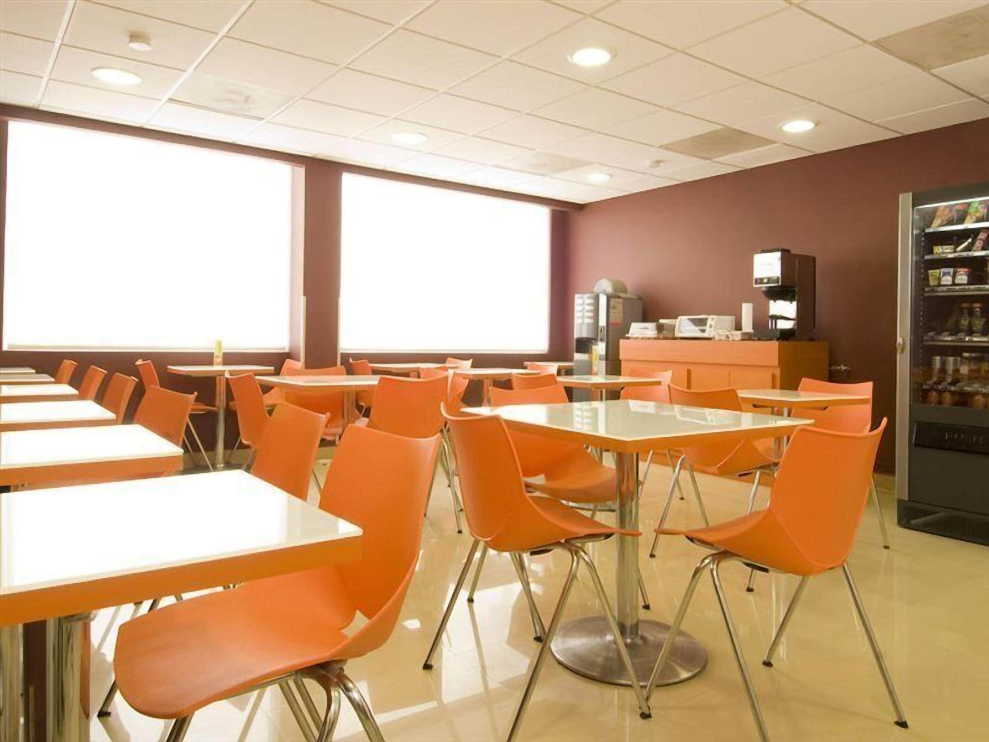 Una cafetería con sillas y mesas naranjas, una máquina expendedora y una estación de café.