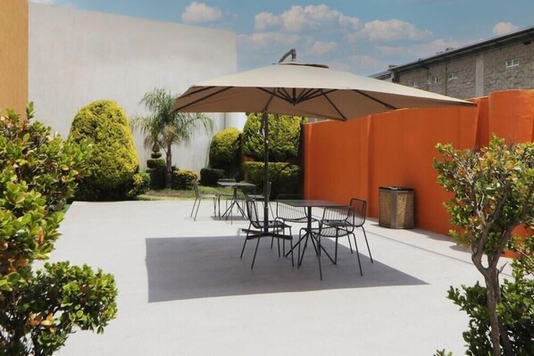 An outdoor patio with a table and chairs under a large umbrella, surrounded by orange walls and greenery. - City Express Junior by Marriott Toluca Aeropuerto (Toluca)