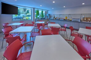 Un restaurante con sillas rojas y mesas blancas, una zona de cocina y amplias ventanas con vistas a la calle.
