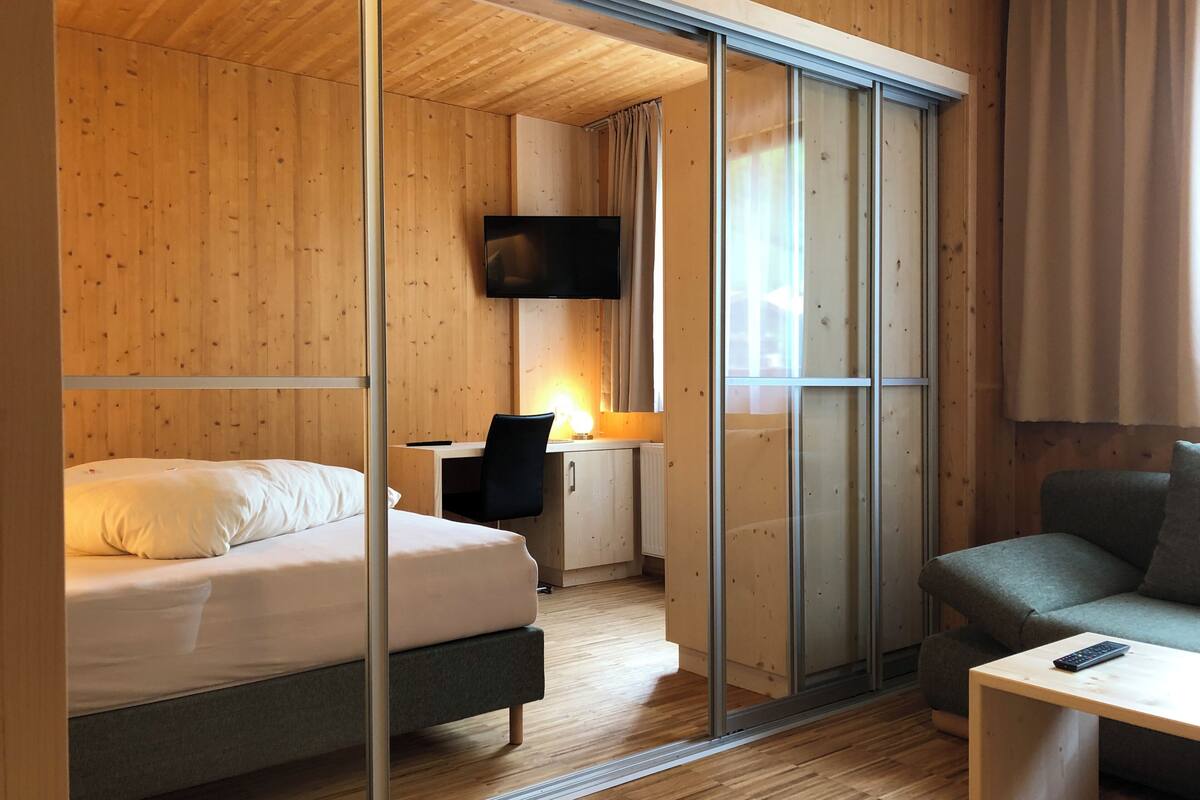 A modern hotel room with a wooden interior, a bed, a desk with a chair, a television, and a sofa.