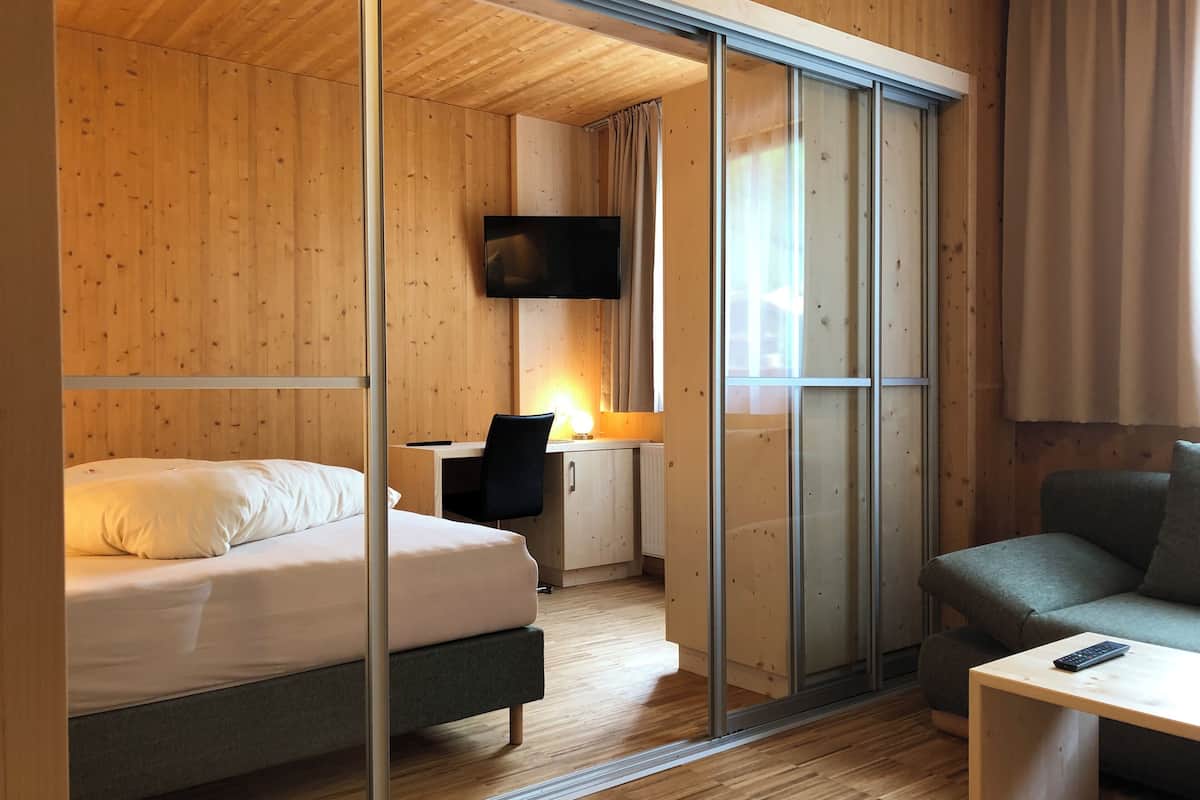 A modern hotel room with a wooden interior, a bed, a desk with a chair, a television, and a sofa.