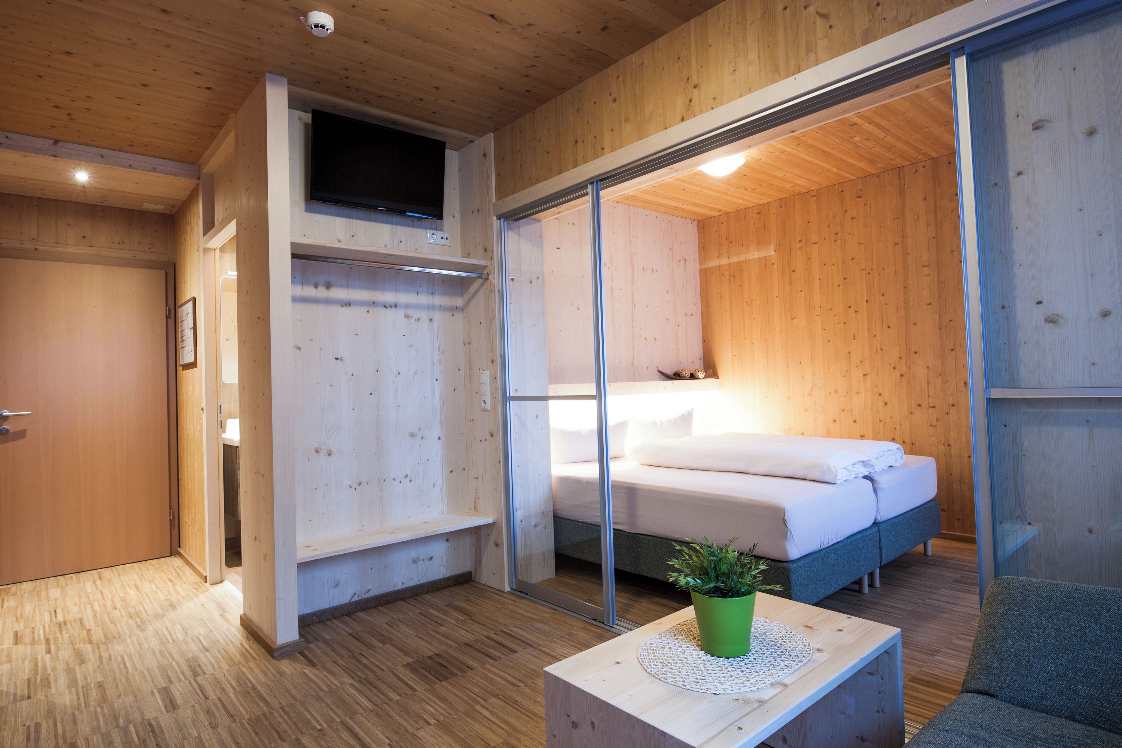 Un dormitorio moderno con suelo de madera, una cama con ropa de cama blanca, una planta verde sobre una mesa de madera y un ventanal corredero que da a otra habitación.