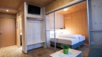 Un dormitorio moderno con suelo de madera, una cama con ropa de cama blanca, una planta verde sobre una mesa de madera y un ventanal corredero que da a otra habitación.