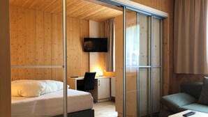 A modern hotel room with a wooden interior, a bed, a desk with a chair, a television, and a sofa.