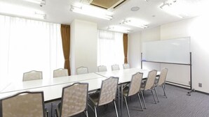 Sala de conferência com uma mesa grande, cadeiras, quadro branco e janelas amplas com cortinas.