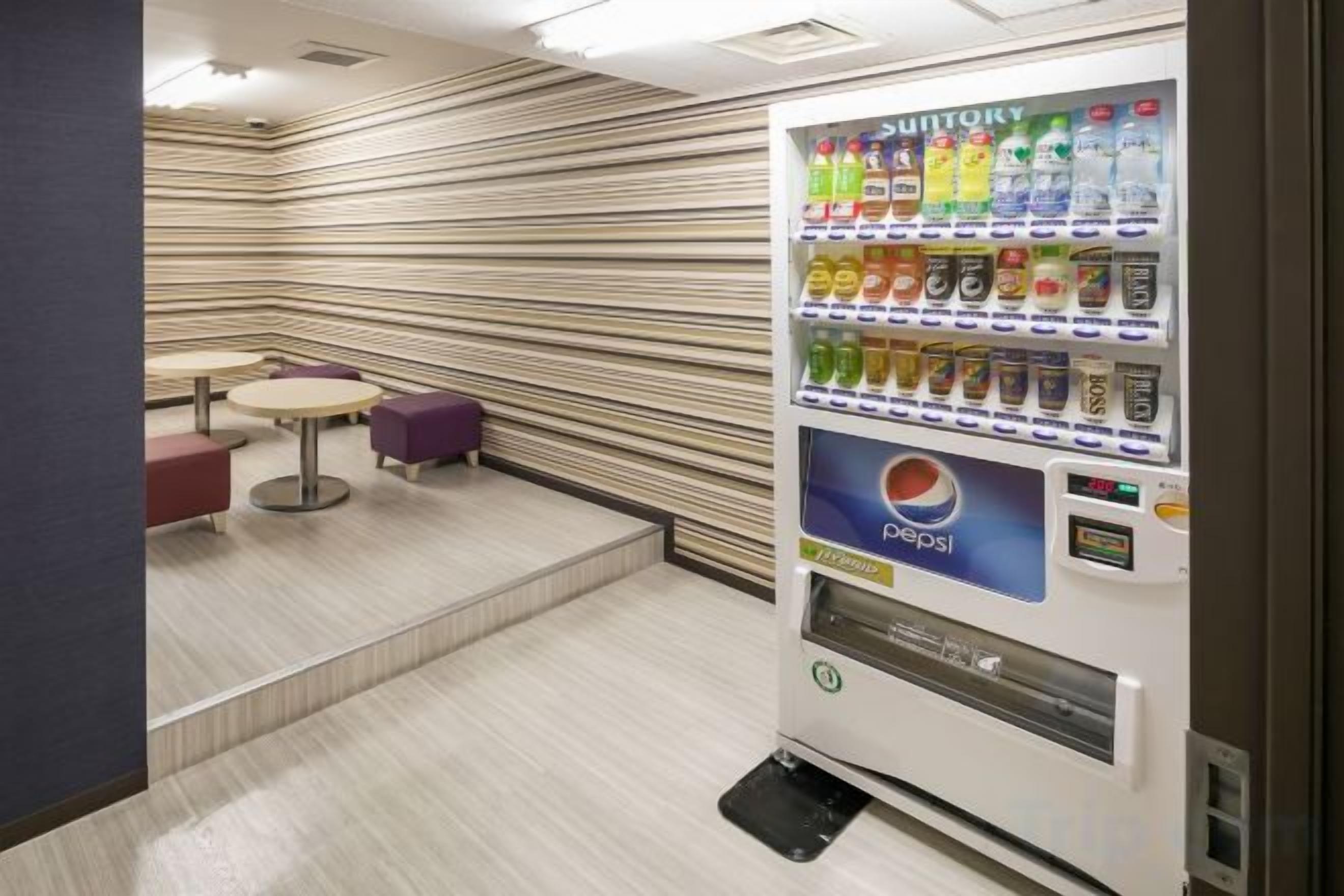 Una macchina distributrice con diverse bevande e il logo Pepsi, situata in una stanza con pareti a righe e un piccolo tavolo e una sedia.
