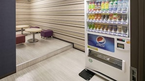 Máquina automática de vendas com o logotipo da Pepsi, rodeada por paredes listradas e uma área de estar com mesas redondas e cadeiras roxas.