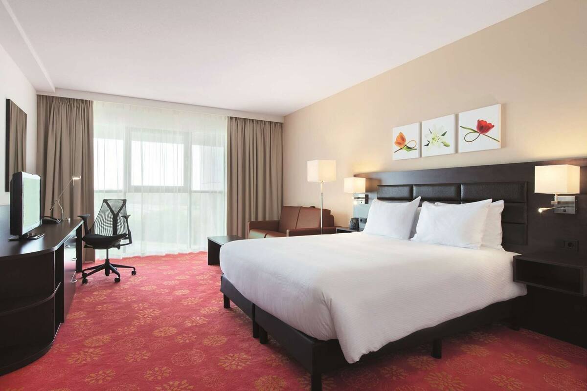 Quarto de hotel com uma cama grande, uma mesa com computador, uma cadeira, uma luminária e obras de arte na parede.
