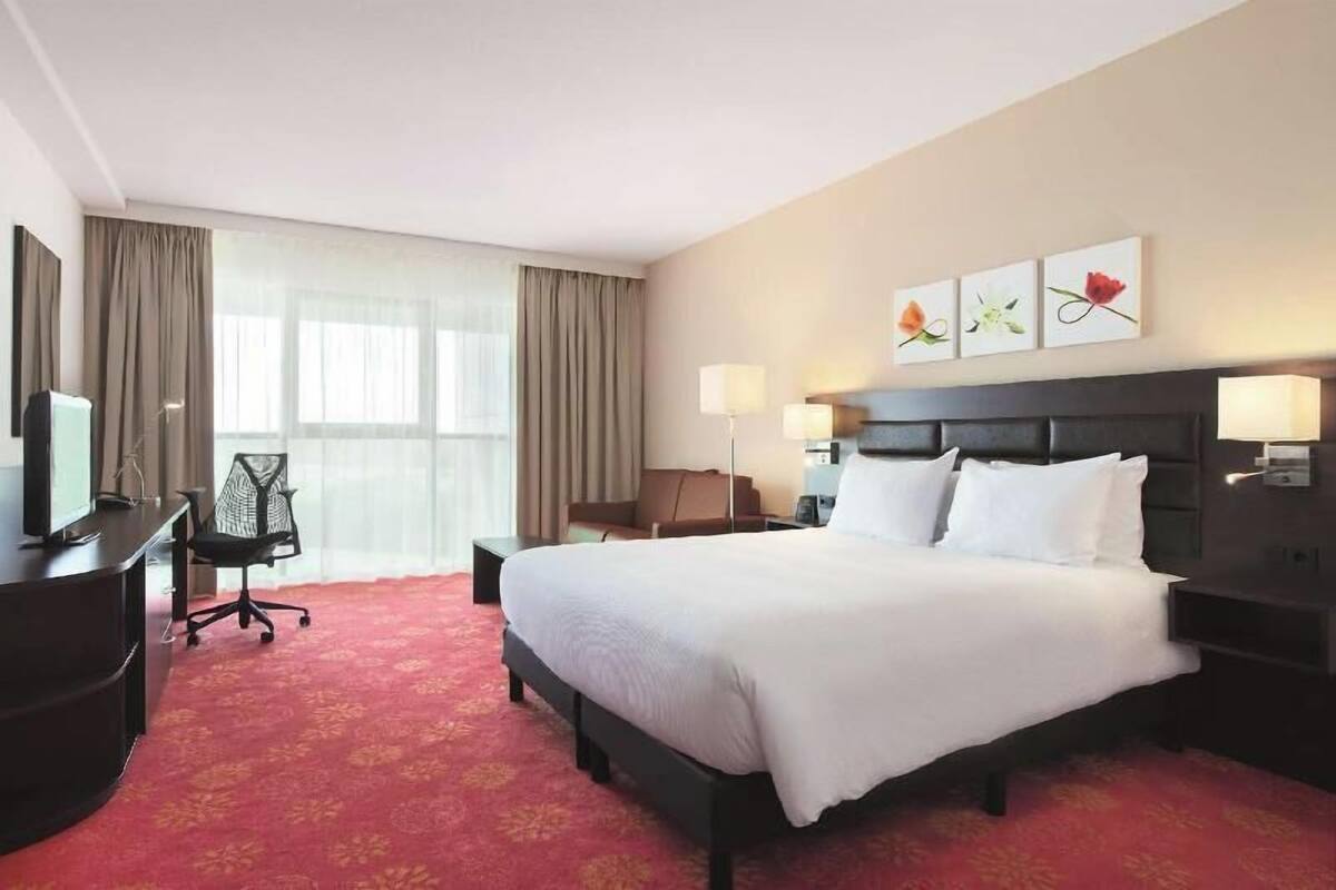 Quarto de hotel com uma cama grande, uma mesa com cadeira, televisão e janela com cortinas.