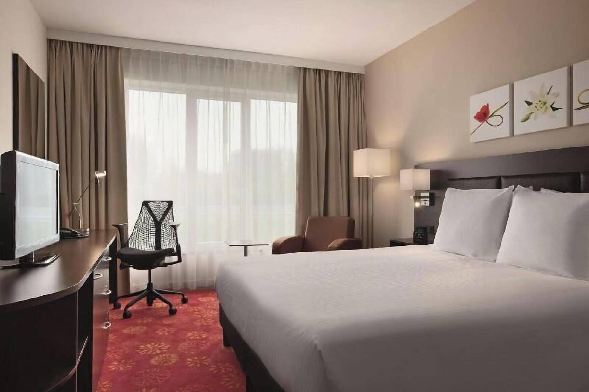 Quarto de hotel com uma cama grande, uma mesa de trabalho com televisão, uma cadeira e uma janela com cortinas.