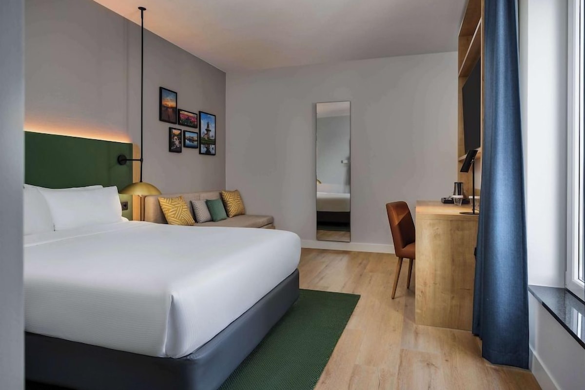 A modern hotel room with a large bed, a desk, a chair, and a mirror.