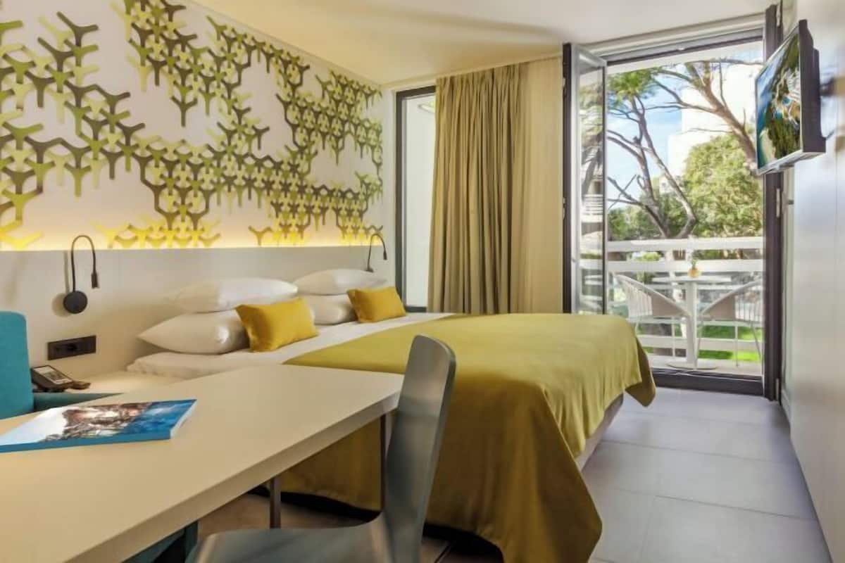 A hotel room with a bed, a desk, and a large window with a view of a balcony and trees.