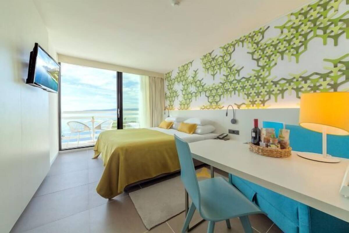 A modern hotel room with a bed, a desk, a chair, a TV, and a balcony with a view.