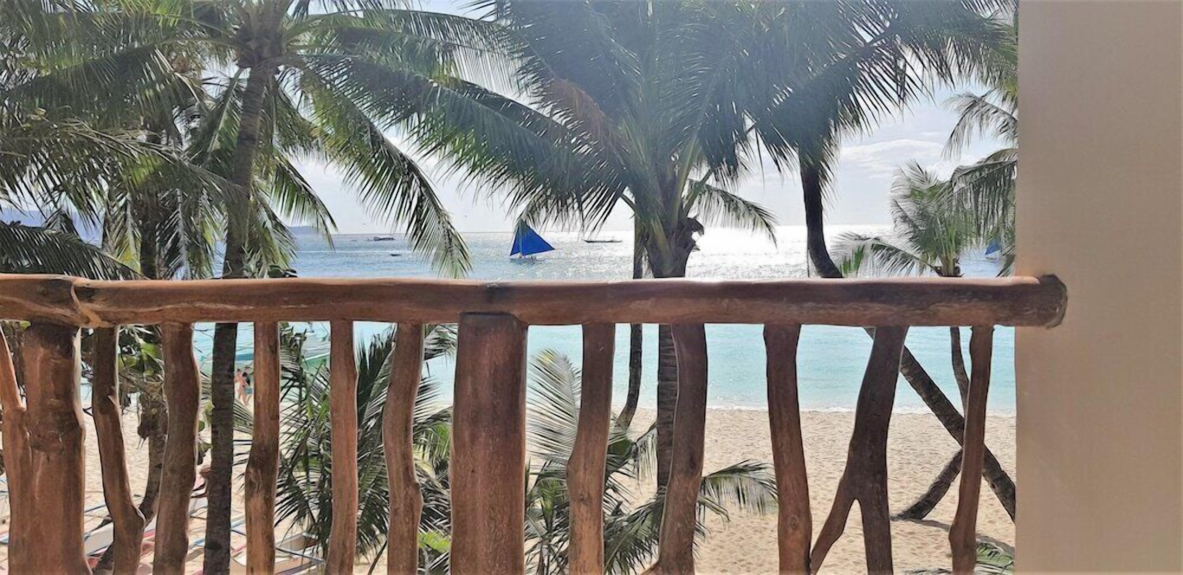 A wooden balcony railing overlooking a beach with palm trees and a sailboat in the distance.