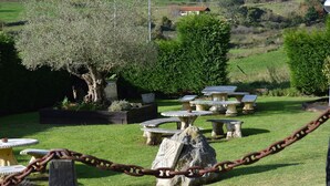 Mesas y sillas de piedra al aire libre en un jardín, con un árbol grande y una casa al fondo.