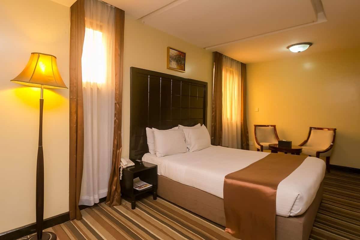 A hotel room with a bed, a nightstand, a lamp, two chairs, and a painting on the wall.
