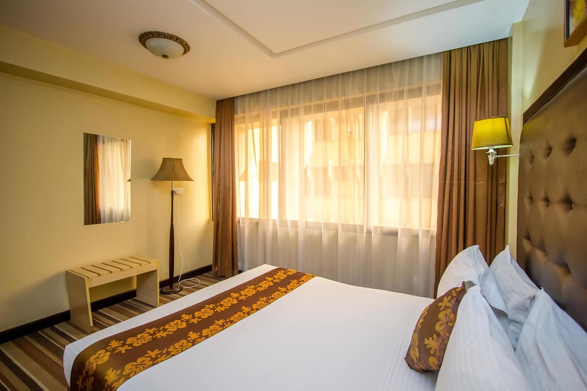 A hotel room with a bed, a bedside lamp, a bench, and a large window with curtains.