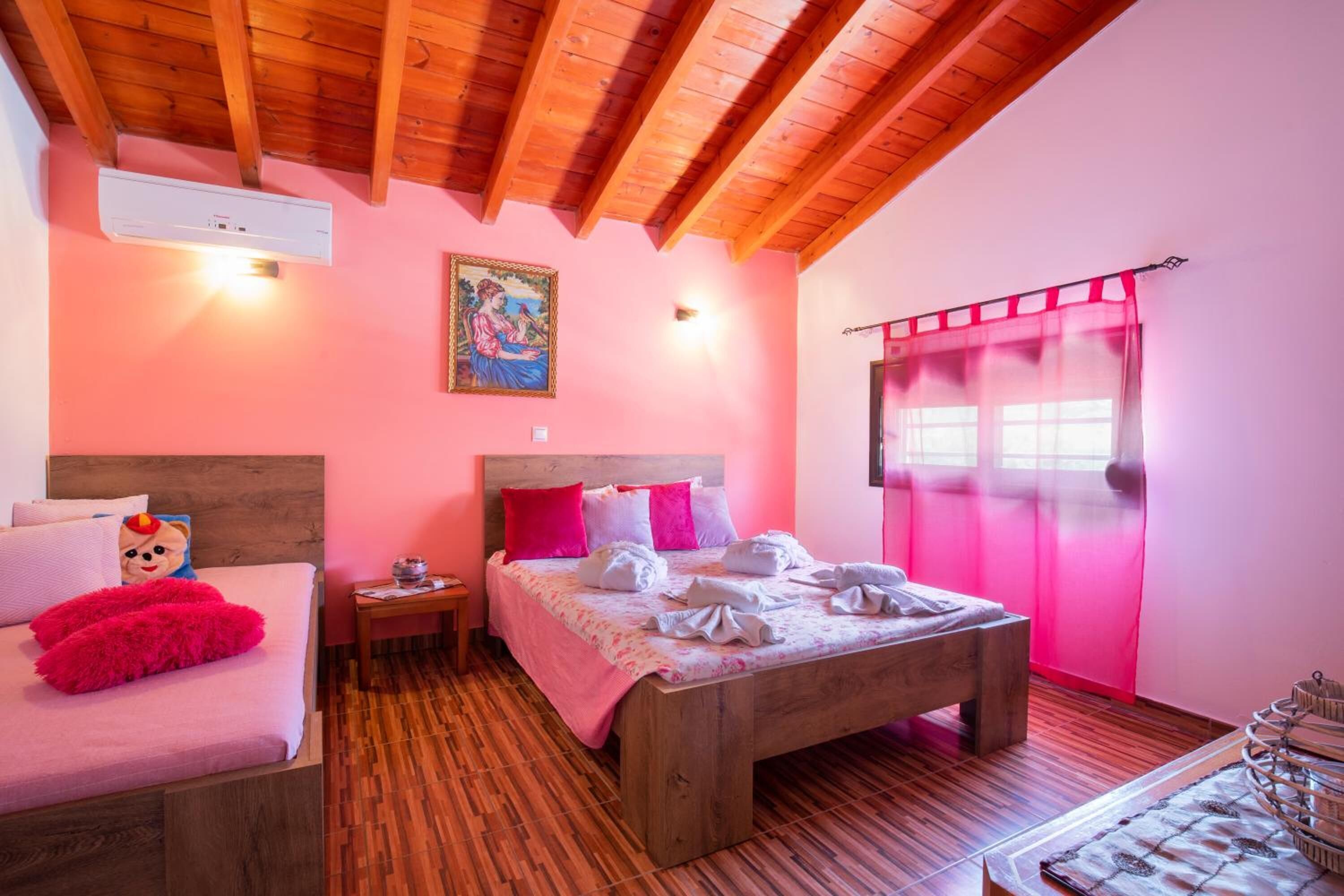 A bedroom with a wooden ceiling, a bed with pink bedding, a nightstand, and a window with pink curtains.