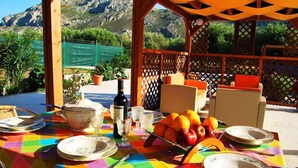 An outdoor dining area with a table set for two, featuring a colorful tablecloth, plates, cutlery, a wine bottle, and a fruit basket. - Stergios Villa (Rhodes)
