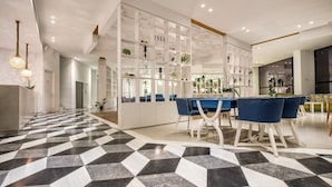 Ein modernes Interieur mit einem Schachbrettmuster-Boden, einer zentralen Essecke mit blauen Stühlen und einer weißen Regaleinheit mit Pflanzen.