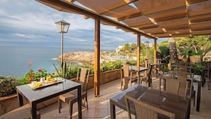 An outdoor patio with wicker furniture, a table with a plate of food, and a view of the sea. - Eden Roc (Sant Feliu de Guixols)