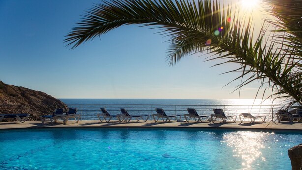A clear blue pool with sun reflecting on the water, surrounded by lounge chairs facing the ocean. - Eden Roc (Sant Feliu de Guixols)