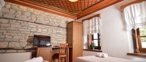 Une pièce avec un mur en pierre, un bureau en bois, une télévision, deux lits, une chaise et une fenêtre avec des rideaux.
