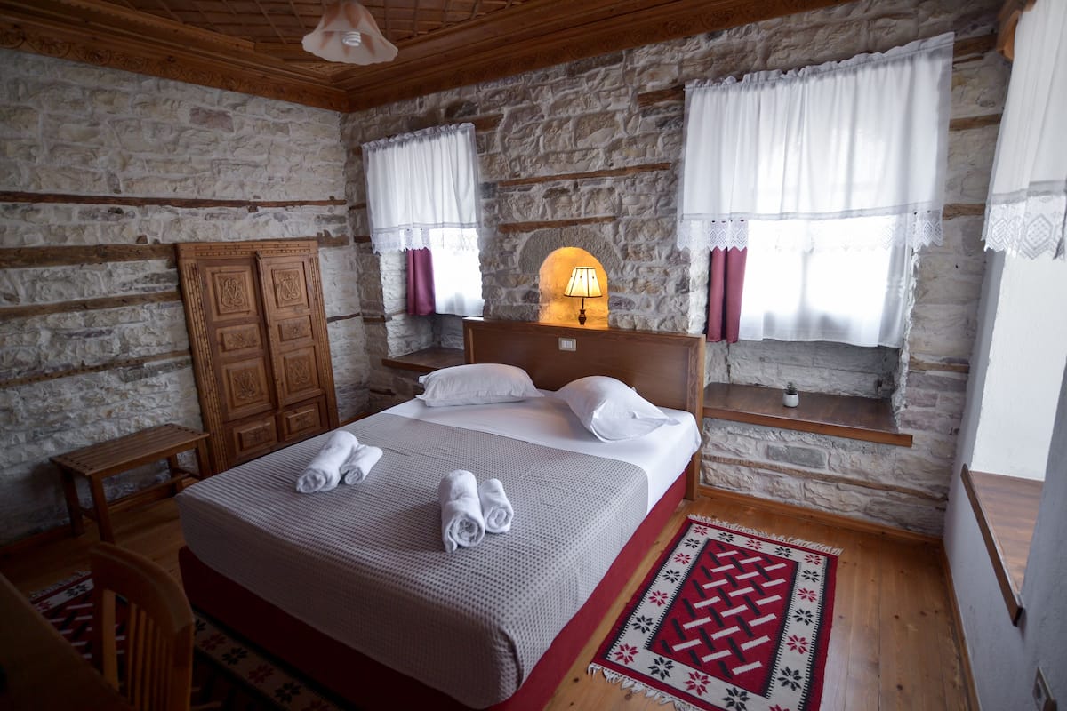 A bedroom with a stone wall, wooden bed, white curtains, and a red patterned rug.