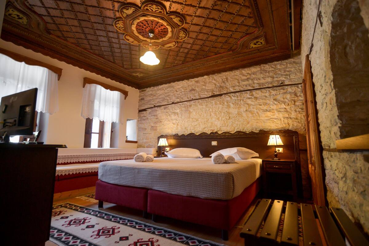 A hotel room with a large bed, a wooden headboard, a stone wall, and a ceiling with intricate designs.