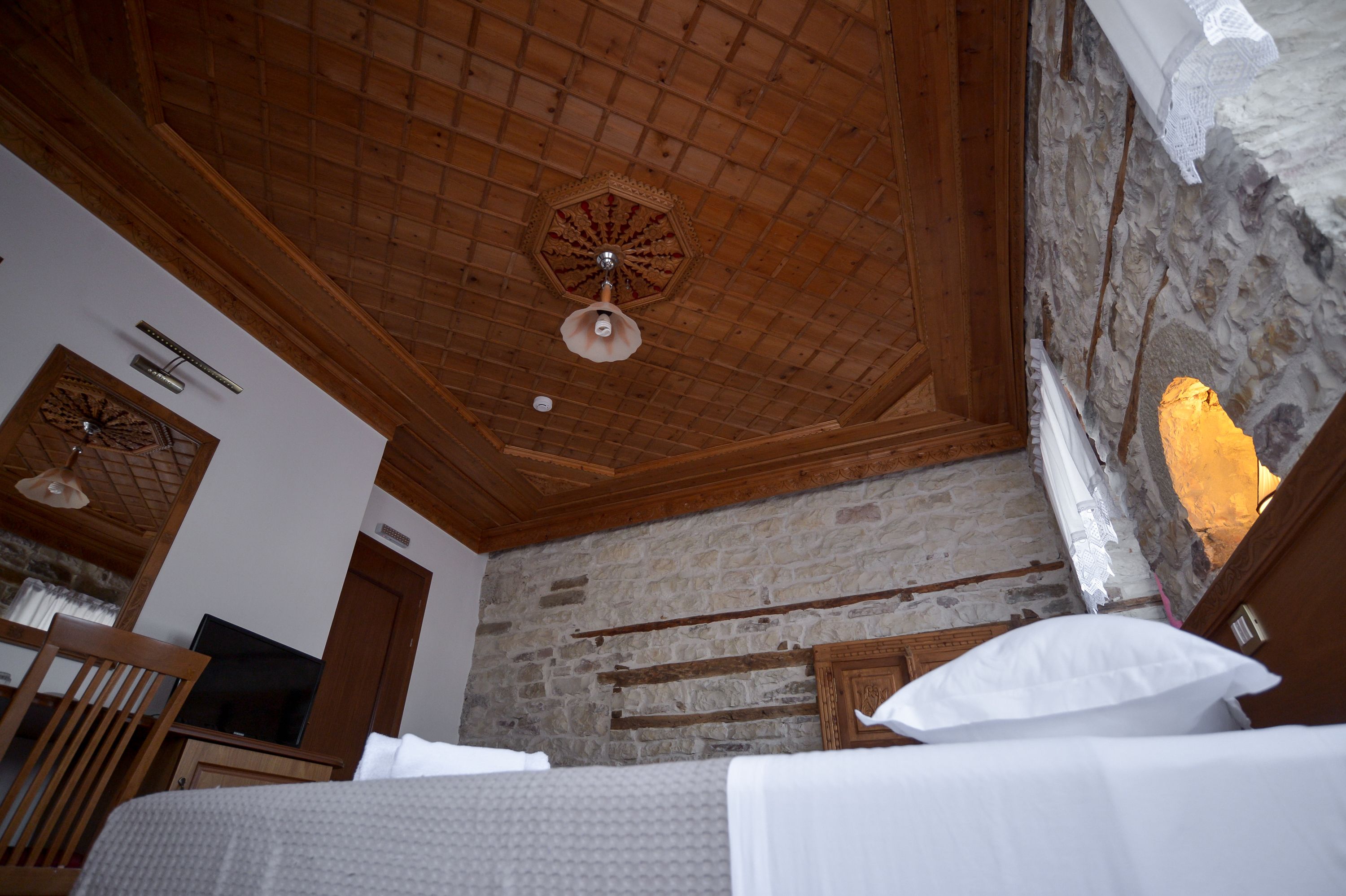 A room with a wooden ceiling, stone walls, a bed, a chair, and a television.