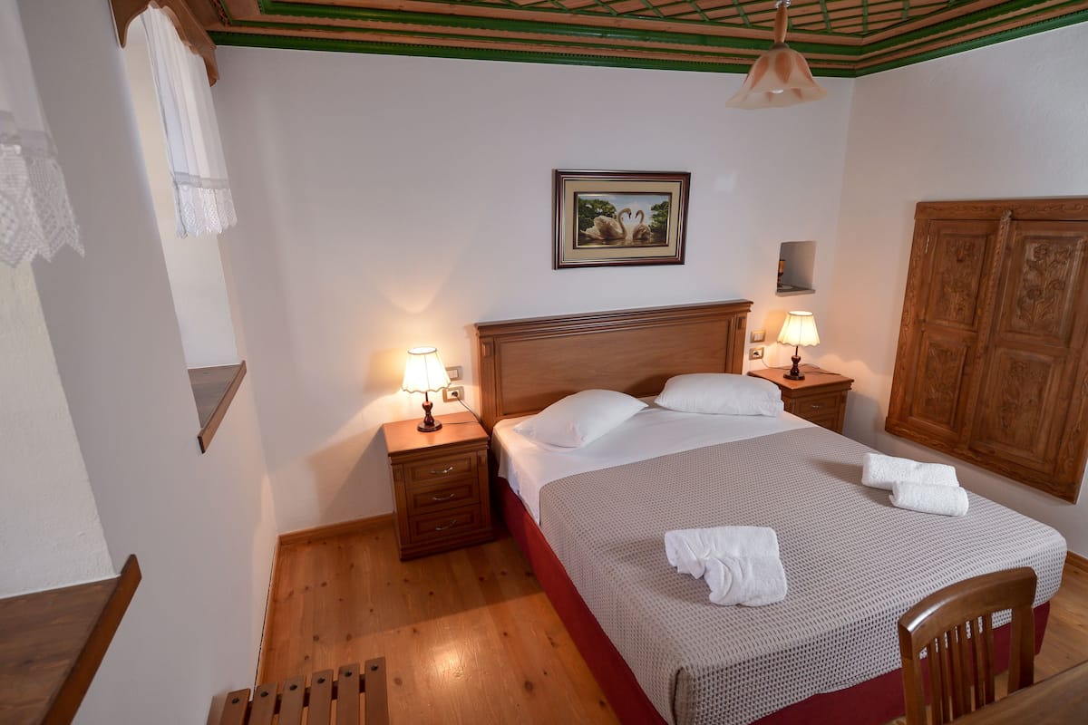 A neatly made bed with white linens and two pillows, flanked by wooden nightstands with lamps, in a room with wooden flooring and a green and white ceiling.