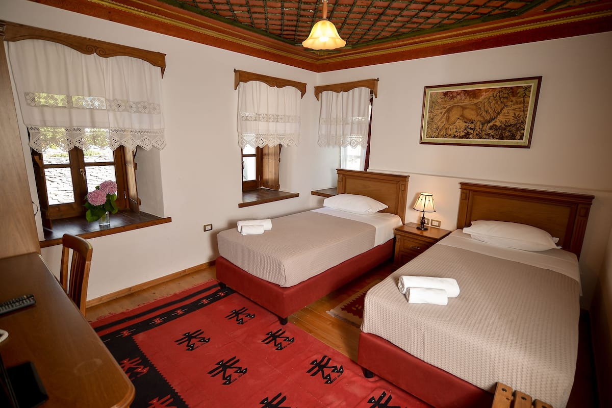 A hotel room with two beds, a wooden desk, a chair, a window with lace curtains, a painting on the wall, and a red patterned rug.