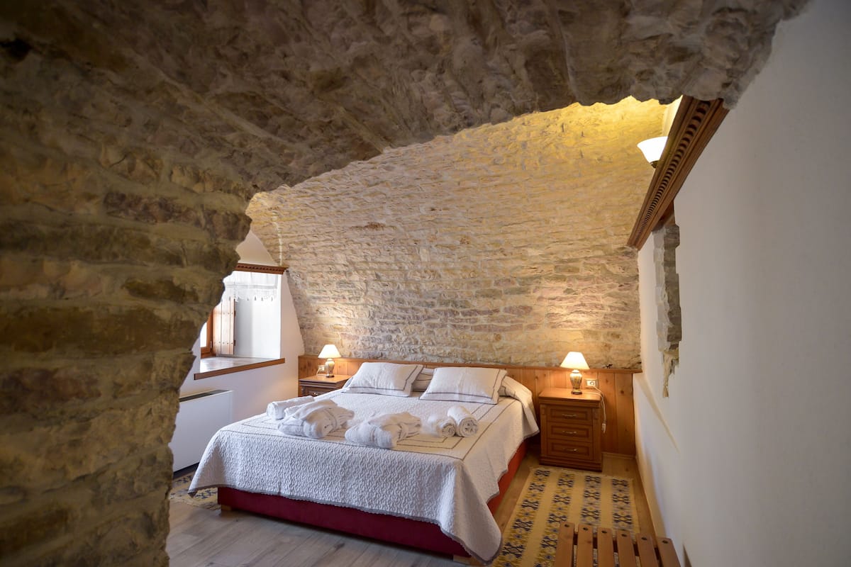 A bedroom with a stone ceiling, a wooden bed, two bedside tables with lamps, and a view of the outside through a window.