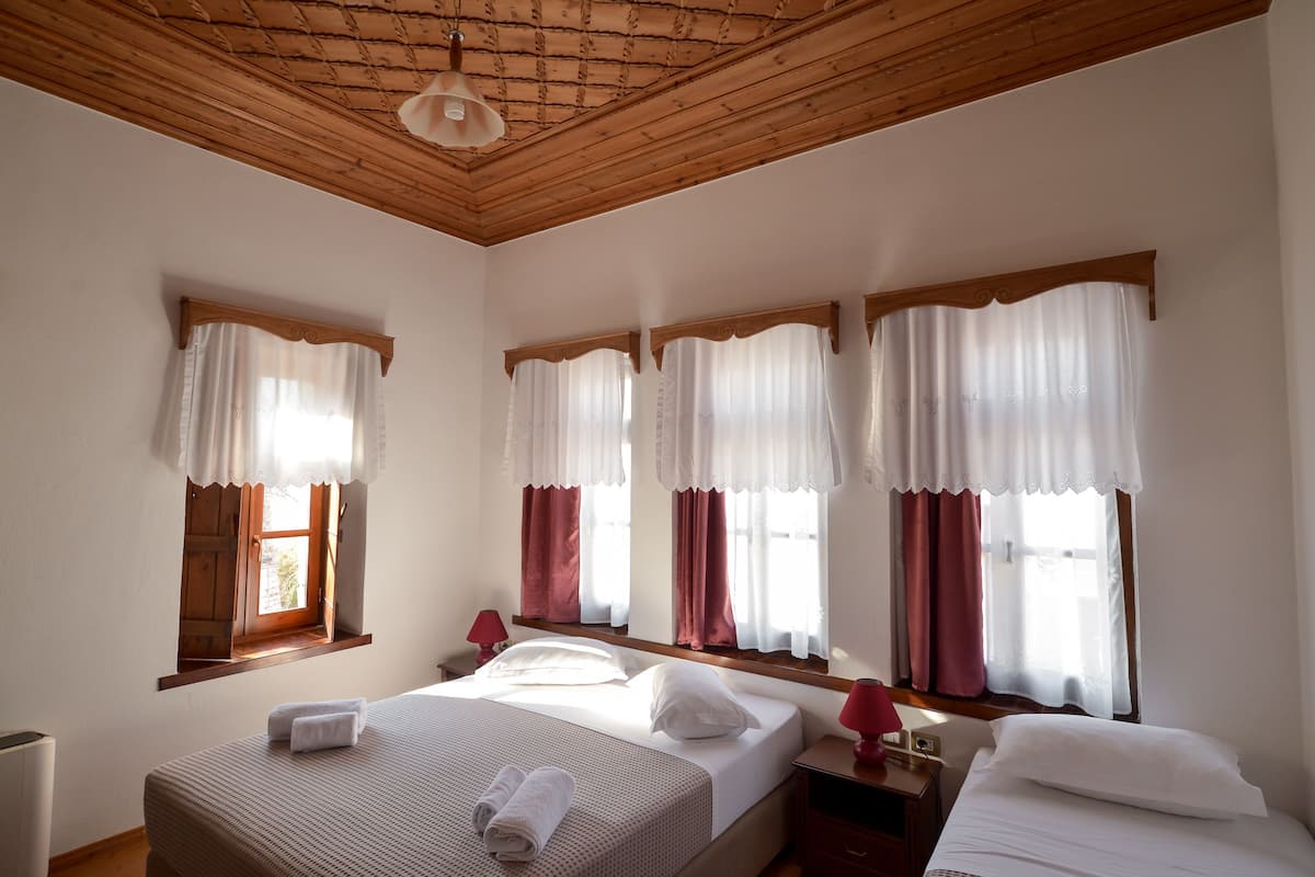 A hotel room with a wooden ceiling, three windows with curtains, a bed with white linens, and a bedside table with a lamp.